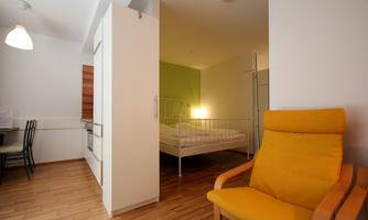 A bright room with a yellow chair and a bed behind a semi-transparent partition. The room features wooden flooring and a relaxed atmosphere.