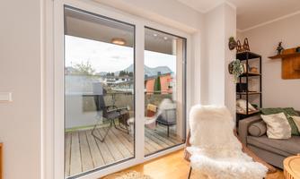 A cozy living space with large windows overlooking a balcony with a wooden terrace. The decor appears modern and inviting.