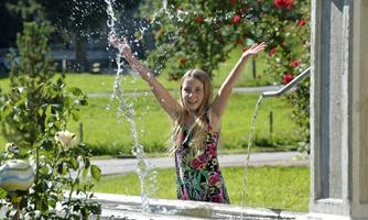 A cheerful girl is playing in the water with her arms raised. In the background, green meadows and roses can be seen.