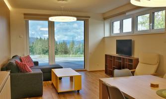 Cozy living room with a gray sofa, a coffee table, and a television. Large windows offer a view of the green landscape outside.