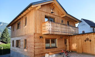A charming wooden house with a balcony and a low fence. It is situated in a rural setting with mountains in the background.