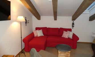 A cozy living space with a red sofa and two cushions. The walls are light, and there are wooden ceiling beams as well as a lamp and a television.