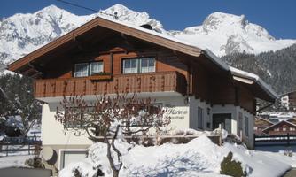A beautiful wooden house in the snow, surrounded by snow-covered mountains. The clear blue sky creates an idyllic winter scene.