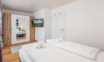 A modern bedroom with a double bed and elegant bedding. In the background, there is a wardrobe, a television, and a window that provides plenty of light.