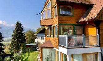A modern wooden house in nature with large windows and a balcony. The garden is green and well-maintained, with trees and mountains visible in the background.