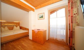 A cozy bedroom with wood paneling and a large window. There is a double bed and a small table with a tablecloth.