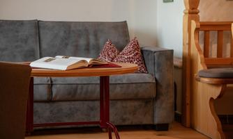 A cozy living room with a gray sofa and a small table. On the table lies an open book and decorative cushions are visible.