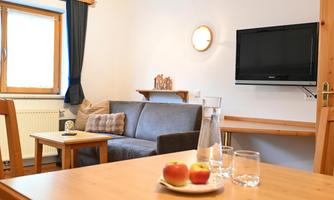 A cozy living space with a gray sofa and a table. On the wall, there is a television, and on the table, there are apples and glasses.