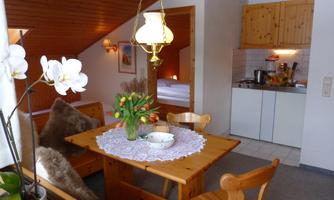 A cozy dining area with a wooden table, fresh flowers, and a warm lamp. In the background, a small kitchenette is visible.