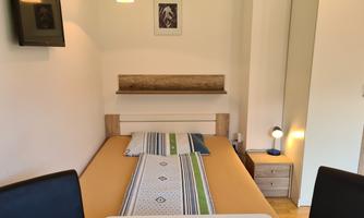 A simple bedroom with a bed, a small table, and a TV. The walls are light, and there is a wardrobe as well as a picture on the wall.