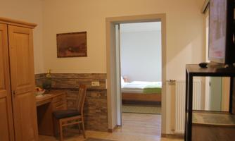 A cozy room with a desk and a wooden cabinet. In the background, a bedroom with a bed is visible.