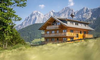 A modern wooden house in the mountains, surrounded by green meadows. In the background, impressive mountain peaks can be seen.