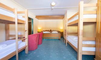 A cozy dormitory with two bunk beds and a double bed. The walls are light, and there are soft seating options in the corner.