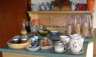 A breakfast buffet with various jams, muesli, and drinks on a wooden cabinet. Flowers decorate the background.
