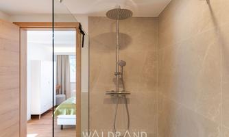 A modern shower cabin with a rain shower-like head. In the background, a bright bedroom with a green bed is visible.