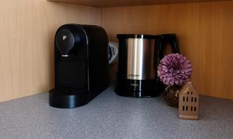 A modern kitchenette with a black coffee machine and a silver kettle. In addition, there is a purple flower and a small decorative apartment in the background.