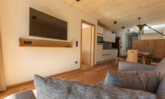 A modern living room with a large television on the wall and a cozy sofa. The room features warm wooden tones and an open access to the kitchen.