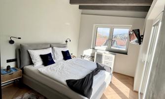 A modern bedroom with a large bed, surrounded by bright walls and a wooden beam ceiling. There is a window with a view and a small work desk.