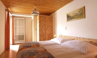 A cozy bedroom with a double bed and wooden furniture. Light streams through the windows, creating an inviting atmosphere.