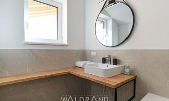 A modern bathroom with a wooden vanity and a round mirror. There is a small window that lets in a lot of light.