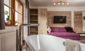 A modern bathroom with a freestanding bathtub and a cozy living area in the background. The wooden details and the large window front create an inviting atmosphere.