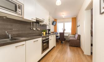 A modern kitchen with white cabinets and wooden flooring. In the background, there is a dining area with a window and light curtains.