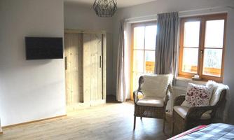 A cozy room with a light wooden floor. There are windows with curtains, two chairs, and a television on the wall.