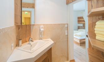 A modern bathroom with wooden furniture and a glass sink. There is a mirror and a view of an adjacent bedroom.