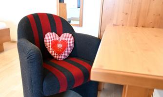 A comfortable chair with a red-black striped cover and a heart-shaped cushion. Next to it stands a wooden table in a bright room.