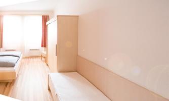 A simple room with wooden flooring and large windows. There are two beds and a bright, plain furnishing.