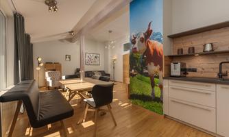 A modern kitchen with a dining area and a large window. On the wall, there is a picture of a cow in a rural landscape.