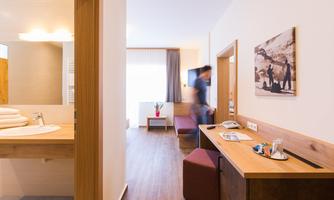 A modern hotel room with a desk and a seating area. The room is bright and cheerfully designed, with a window that lets in daylight.