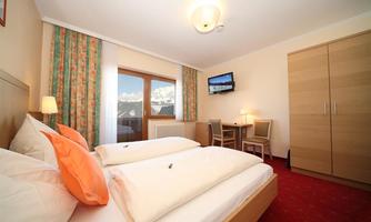 A cozy hotel room with two beds, a table, and a television. The window offers a beautiful view of the mountains.