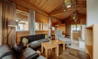 A cozy living area with wooden furniture and large windows. The room is brightly lit and invites relaxation.
