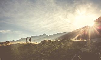Two people are standing on a hill and looking at the landscape in the sunlight. In the background, there are mountains and a clear sky.