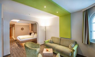 A modern room with a cozy seating area and a double bed in the background. The walls are painted in light colors, with a striking green accent.