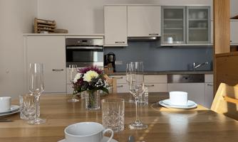 A modern kitchen with a wooden table set with dishes and glasses. In the center, there is a bouquet of flowers on the table.