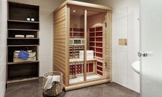 A modern infrared sauna with a glass front and wooden paneling. Next to it are shelves with care products and towels.