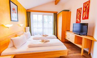 A cozy bedroom with two beds, warm wooden furniture, and colorful wall murals. Large windows ensure plenty of natural light.