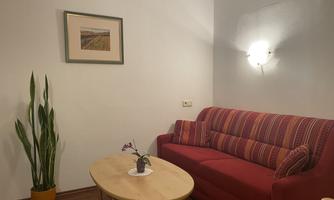 A cozy living room with a red sofa and a round table. A picture hangs on the wall, and a plant stands in the background.