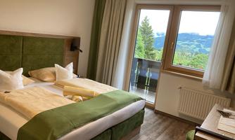 A cozy hotel room with a large double bed and green decorative elements. The view from the window shows a beautiful landscape with hills and trees.