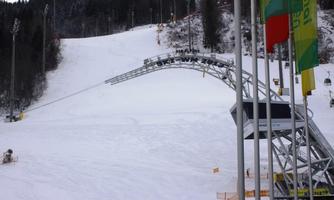 A snow-covered ski slope with a ski lift in the background. The surroundings are surrounded by trees, and the slope is well-groomed.