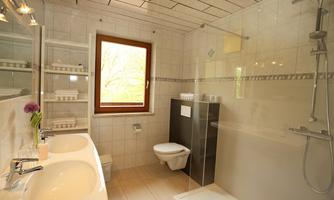 A modern bathroom with two sinks and a glass shower. A window lets in daylight and there is an elegant, bright design.