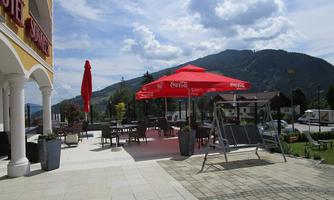 A cozy terrace with red umbrellas and seating. In the background, gentle hills and a blue sky with clouds can be seen.