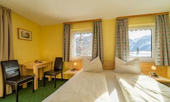 A cozy room with a large bed, two chairs, and a table. Through the windows, one looks out at a picturesque mountain landscape.
