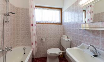 A simple bathroom with a bathtub, a sink, and a toilet. The walls are tiled in light pink, and there is a window with curtains.