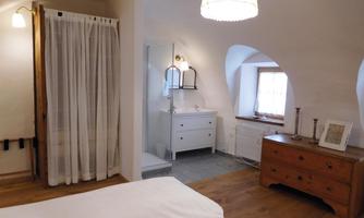 A cozy bedroom with a large window and warm wooden floors. The decor is modern and bright, with a bathroom area nearby.