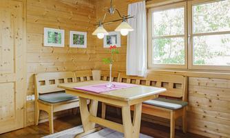A cozy dining area in a wooden house with a table and benches. There is a flower vase on the table and bright windows that let in light.