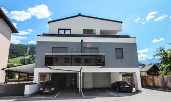 A modern multi-story building with a flat roof. It has parking spaces in front of the entrance and is located in a picturesque setting.