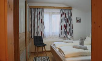 A cozy bedroom with two beds and a chair. The windows are adorned with curtains and the room has a warm wooden character.